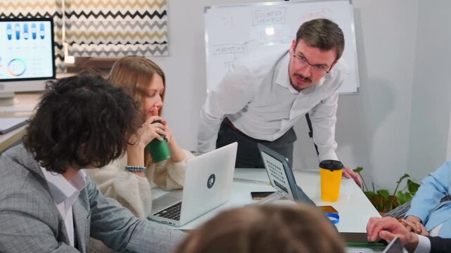 Authoritative male manager stands over table, sternly addressing employees about poor performance and possible layoffs. Colleagues listen seriously, some avoiding eye contact, during a tense meeting.
