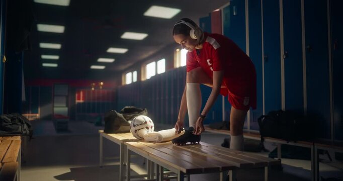 Woman Athlete in Red Soccer Uniform Preparing for a Match a Locker Room, Listening to Music on Headphones as She is Tying Laces on Her Cleats. Female Footballer Gathering Focus Before Training