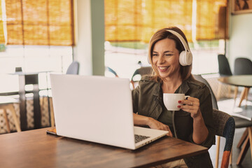 Woman in headphones laughing with coffee while working on a laptop in a cafe, joining a video call and enjoying flexible remote work, connectivity and casual entrepreneurship