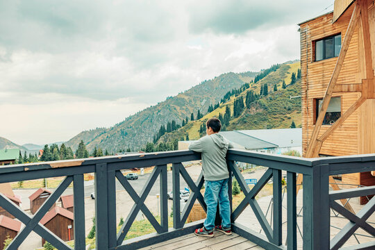 A young Asian man stands on a wooden balcony overlooking green mountains. The scene captures a tranquil moment in a recreational travel setting.