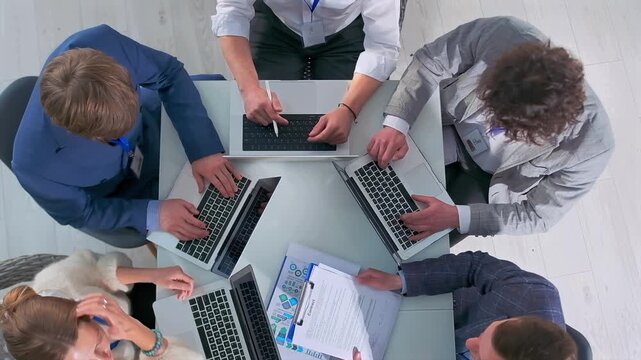 Top view of group of office employees collaborating at meeting table. Team members type on laptops and review documents, engaged in a corporate work process. Teamwork and professional cooperation.