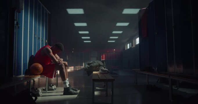 Disappointed Young Basketball Player Sitting in a Locker Room, Head Down, Thinking About the Loss in the Game. Tense Atmosphere Highlights the Emotional Pressure of Professional Sports