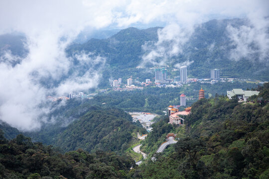 View of a modern city nestled amidst lush green mountains, partially obscured by swirling clouds, contrasting natural beauty with urban development, Genting Highlands, Pahang, Malaysia.