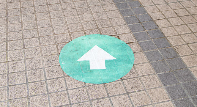 White arrow in circle green on ground square brick floor of sidewalk on side of road. Design elements of directional signs on walkway. Temporary road marking or wayfinding symbol sign. 