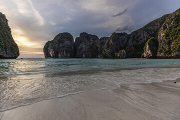 View of turquoise waters of Maya Bay meeting a sandy beach under a dramatic sky, with limestone cliffs rising majestically from the sea, Phi Phi Islands, Krabi, Thailand.