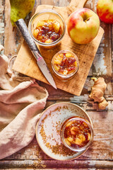 Flat lay of apple chutney in glass jars, surrounded by fresh apples, pears, ginger, and a kitchen knife on a rustic wooden surface. Homemade preserve with seasonal fruit and spices.