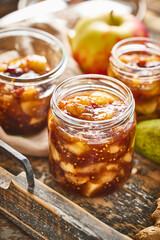 Homemade apple chutney in jars displayed in a rustic wooden tray with fabric and spices. Traditional fruit preserve with ginger, mustard seeds, and seasonal fruit.