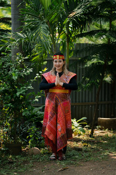 A woman in Batak traditional dress from North Sumatra posing outdoors, wearing ulos fabric with red ethnic patterns and headband, symbol of Indonesian heritage.