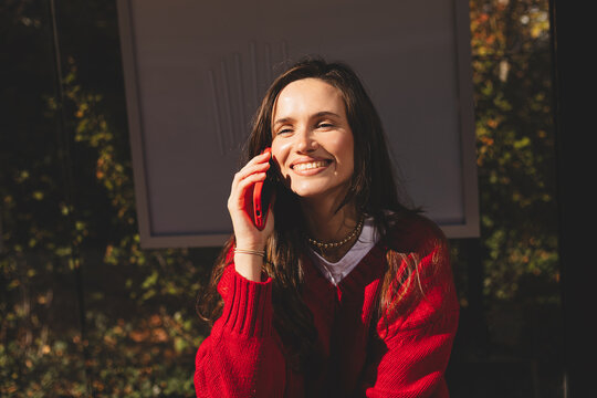 Young tourist waiting at the bus station, chatting on her smartphone while enjoying a sunny day in the vibrant city atmosphere. Woman wear red sweater, laughing and smiling. - Powered by Adobe