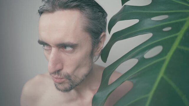 Curious Man with Striking Blue Eyes Transitions from Intrigued to Contemplative Behind a Large Green Monstera Leaf