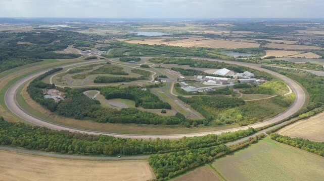 Aerial view of the Millbrook Proving Ground test track surrounded by green trees and golden fields under a cloudy sky, Bedford, England, United Kingdom.