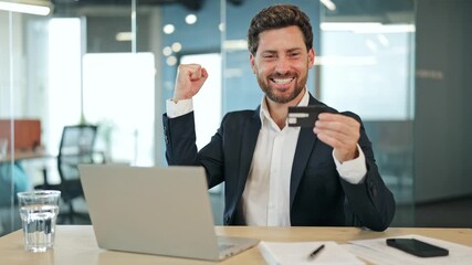 Excited professional businessman, banker, credit officer in a suit jacket triumphantly celebrates a successful online payment holding a credit card at his modern office desk.