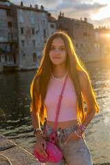 Teen girl posing by canal at sunset. Stylish teenage girl in pink outfit posing near waterfront at...