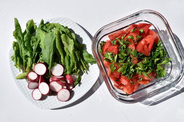 Salad made of red tomatoes and greens in a square glass dish and a plate with radish slices and lettuce leaves