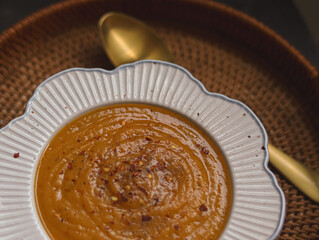 Creamy bowl of pumpkin or butternut squash soup topped with cracked pepper and chilli flakes, placed beside golden spoon on woven mat, concept of homemade seasonal food in the English kitchen