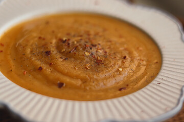 Creamy bowl of pumpkin or butternut squash soup topped with cracked pepper and chilli flakes, placed beside golden spoon on woven mat, concept of homemade seasonal food in the English kitchen