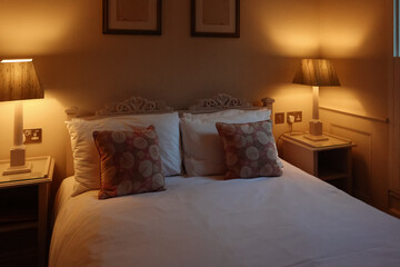 Evening view of traditional English bedroom with lit bedside lamp, carved headboard, floral cushions and shuttered window, concept of home comfort, quiet rest and warm countryside interior