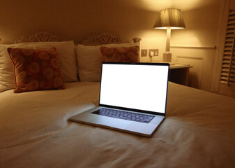 Open laptop mockup with blank white screen on neatly made bed in traditional English bedroom with floral cushions and bedside lamp, concept of work from home, evening routine and digital lifestyle