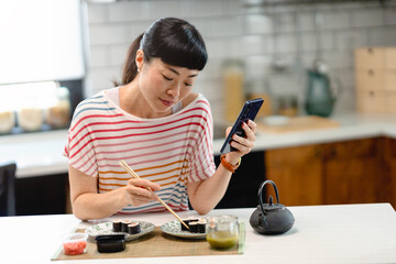 Cheerful Japanese woman eating sushi with chopsticks while looking at smartphone in kitchen. Happy Asian woman enjoying meal, technology and modern home lifestyle