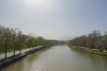 Wide, straight canal or river running between tree-lined roads with distant hazy mountains under a bright, misty sky. Spring landscape scenery.