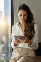 Fototapeta premium A professional woman in a white blouse and beige trousers stands by a large office window, holding and using a tablet