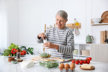 Cooking process. Senior woman adding spices to salad at white marble table in kitchen