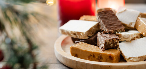 Assorted traditional Christmas nougat on wooden plate and christmas tree background. Copy space