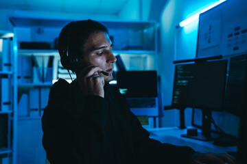 Hacker in black hoodie with headset using computer in dark office, representing cyber crime, data breach, and cybersecurity hacking.