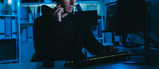 Hacker in black hoodie with headset using computer in dark office, representing cyber crime, data breach, and cybersecurity hacking.