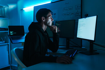 Caucasian male programmer and IT specialist in black hoodie is sitting in dark office, focusing on multiple computer screens, analyzing code, cybersecurity tasks, software development for technology.