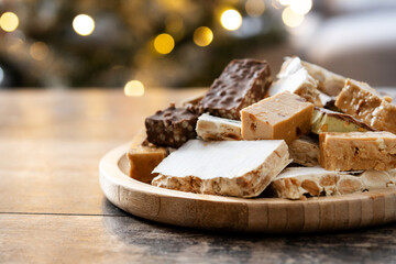 Assorted traditional Christmas nougat on wooden plate and christmas tree background. Copy space