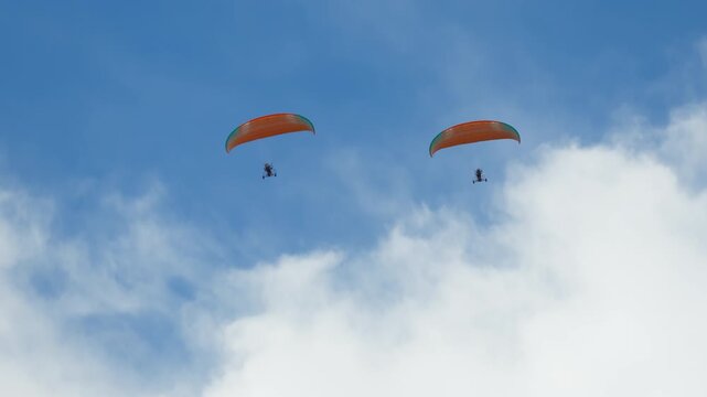Two individual paramotors, each expertly piloted, captured soaring gracefully high altitude against boundless, pristine clear blue sky. paramotors feature distinct orange green canopies, highlighting