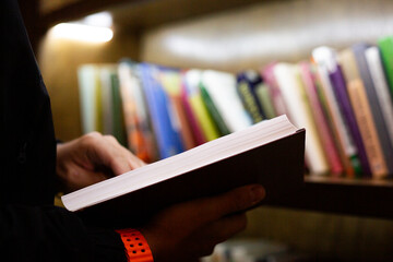 Close up of person holding book in library setting. The photo highlights moment of reading, with shelves full of books in background, creating calm intellectual atmosphere