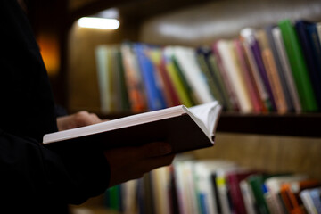 Enjoys reading hardcover book, surrounded by bookshelves in cozy library. Book pages and intellectual ambiance conveys quiet, personal reading experience, ideal for study and relaxation