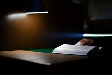 View of open book on desk, with hand gently turning page. Warm, focused light from desk lamp highlights text, creating serene atmosphere of study and concentration in minimalist workspace