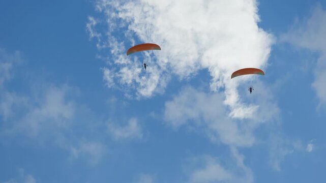 Two paramotors flying against vast, vibrant blue sky adorned with scattered white cumulus clouds. Each motorized paraglider unit, consisting of pilot operating engine propeller system, suspended