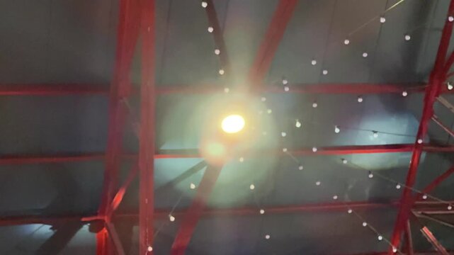 A low-angle shot of a building's ceiling, featuring a large bright light bulb and strings of smaller lights against a backdrop of red metal trusses and rafters