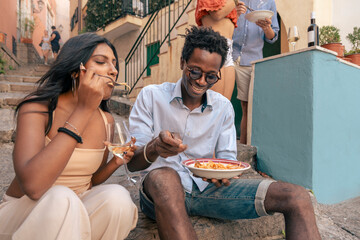 Happy multicultural couple eating pasta on vacation in Italy. Black man and South Asian woman sharing food and wine. Travel and food lifestyle concept.