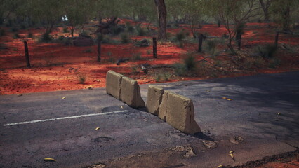A roadblock made of concrete sits on a remote outback road, surrounded by vibrant red earth and sparse vegetation. The midday sun casts shadows, highlighting the rugged beauty of the area.