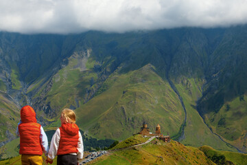 Fototapeta premium Two young girls holding hands and looking at the famous Gergeti Trinity Church in Georgia with many tourists around, capturing a moment of travel, culture, and heritage in the scenic mountains.