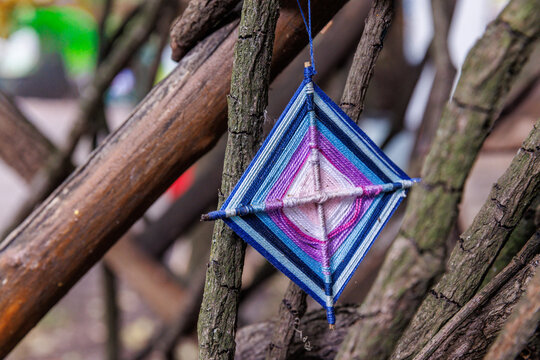 Colorful textile art piece dangling from intertwined branches, showcasing a stunning geometric design with vibrant hues of blue, pink, and white in unique patterns and textures.