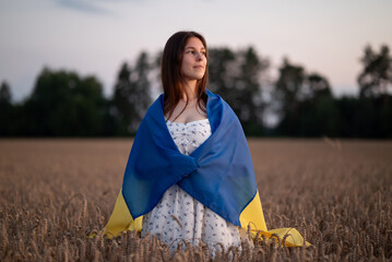 Girl wrapped in Ukraine flag standing in a wheat field at sunset. A young woman in a white dress stands in a ripe wheat field, wrapped in the national flag of Ukraine, a symbol of patriotism and hope