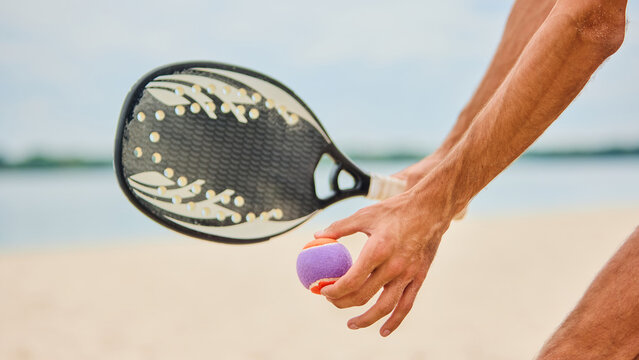Close up of male hand holding paddle racket and small ball on sandy beach. Concept of summer sport, lifestyle, equipment, outdoor leisure and playful emotional anticipation.