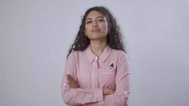 A young South Asian woman with curly brown hair stands confidently, with her arms crossed and a slight smirk