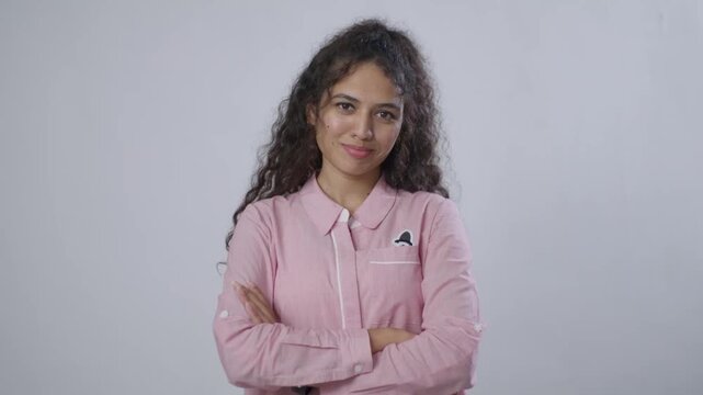 A young South Asian woman with curly brown hair stands confidently, with her arms crossed and a slight smirk