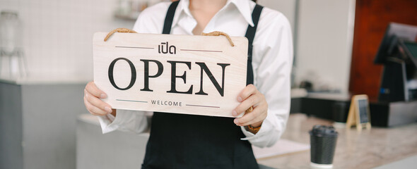 A cafe worker in a black apron and white shirt holds up a wooden "OPEN" sign with Thai text and "WELCOME" written on it. A cup of coffee is on the counter to the side.