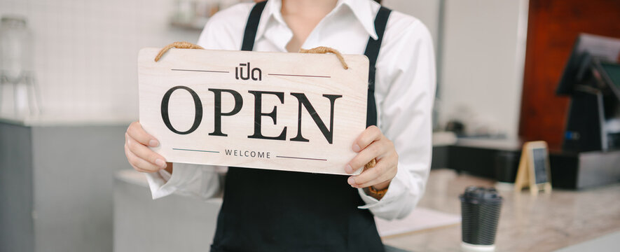 A cafe worker in a black apron and white shirt holds up a wooden "OPEN" sign with Thai text and "WELCOME" written on it. A cup of coffee is on the counter to the side. - Powered by Adobe