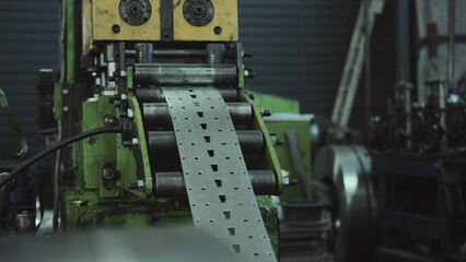 Close up of industrial machinery processing a metal steel strip, forming a repeating pattern of slots and small holes, showcasing manufacturing automation in a workshop setting