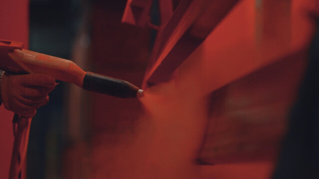 Worker's gloved hand operates an electrostatic spray gun, applying a vibrant red powder coating onto a metal surface in an industrial manufacturing environment, ensuring durable protective finish