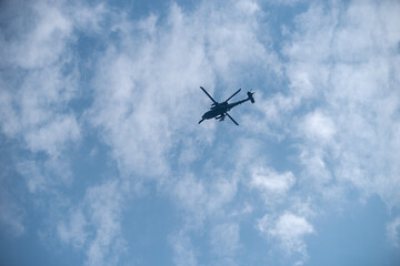 Helikopter Silhouette vor blauem Himmel mit Wolken, Ein Hubschrauber fliegt als dunkle Silhouette vor einem blauen Himmel mit weißen Wolken – dynamische Luftaufnahme in minimalistischem Stil.